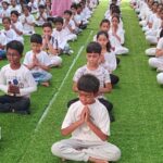 Children in white outfits sit on green grass with eyes closed and hands in prayer, conveying tranquility and focus.
