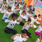 Children in white shirts practice yoga on green grass, focused, with a colorful mural in the background.