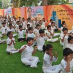 Children in white uniforms meditate cross-legged on grass, with a colorful motivational mural in the peaceful background.