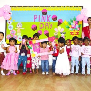 Children and teacher smile in front of a colorful "Pink Day" backdrop, with kids dressed in pink for a joyful celebration.