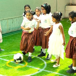 Young girls in school uniforms and a white dress gather on a sunny soccer field, excitedly focused on a soccer ball.