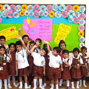 Young children in school uniforms stand cheerfully before a colorful classroom bulletin board with paper flowers and welcome signs.